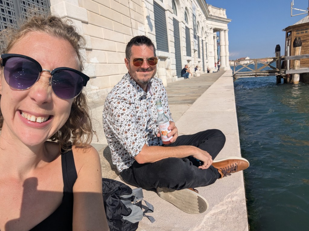 A woman and a man sitting by the water in a sunny outdoor setting. The woman has curly hair and is wearing sunglasses, smiling at the camera, while the man is sitting beside her, holding a beverage and wearing a patterned shirt.