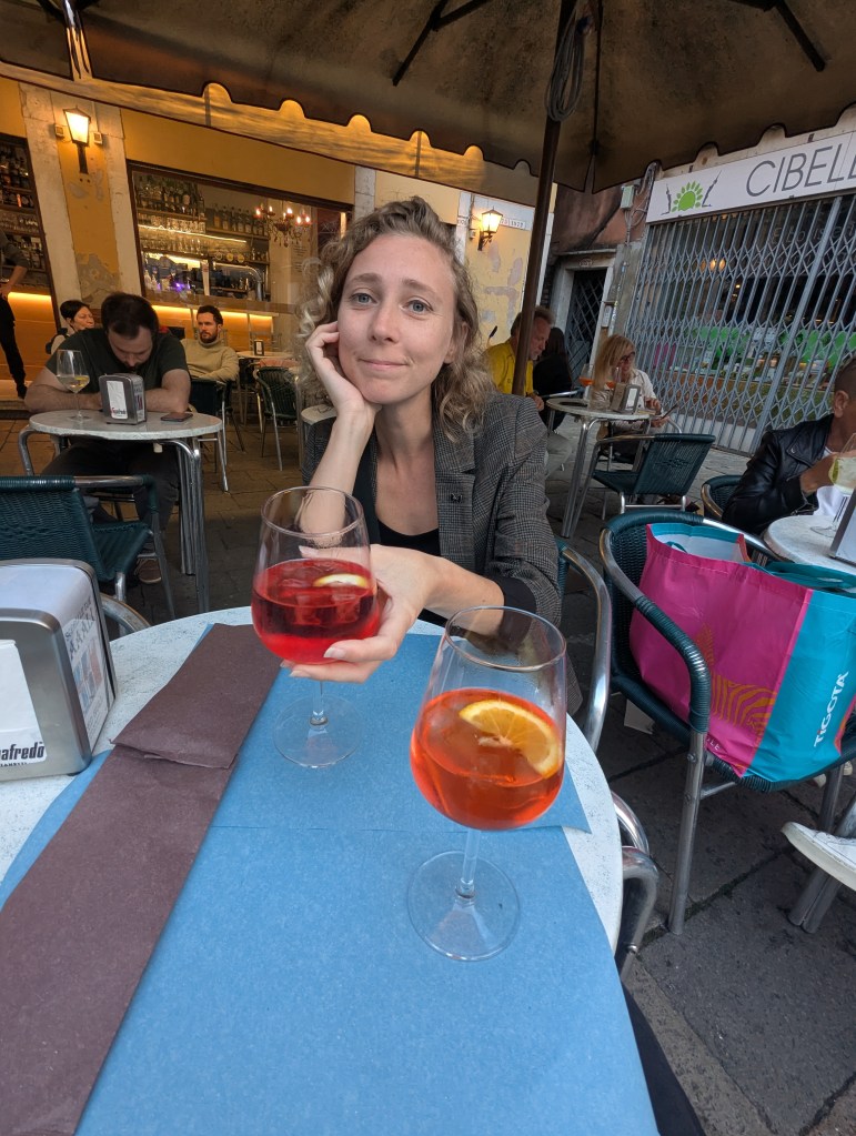 A woman sitting at an outdoor café table, holding a glass of drink with an orange slice, while another glass sits on the table. The background features other patrons and a bar.