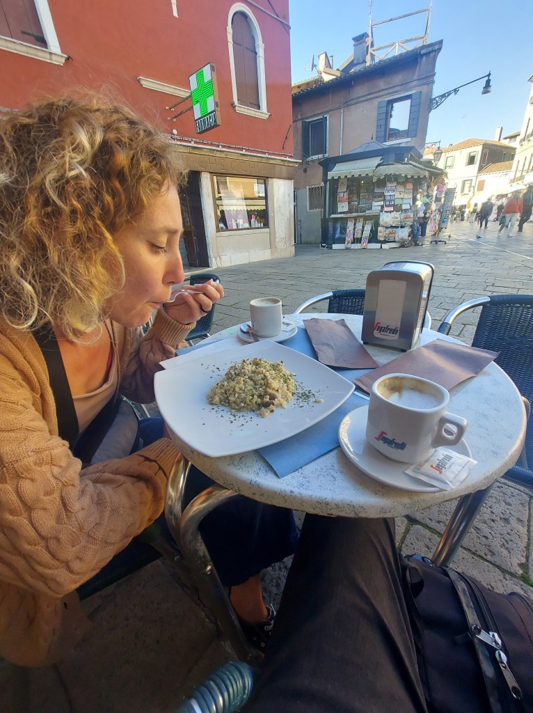 A woman with curly hair enjoying a plate of food at an outdoor café table with a coffee cup beside her. The setting appears to be a lively street in a city with nearby shops.
