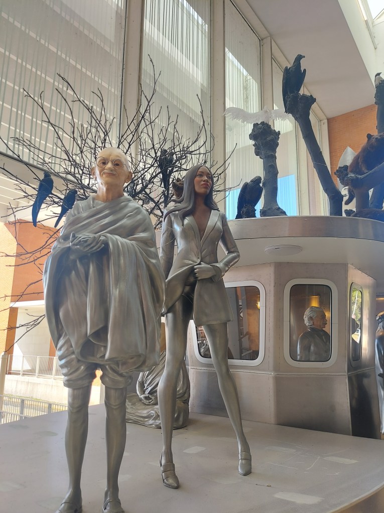 A collection of metallic sculptures depicting two figures, one resembling a monk in traditional attire and the other a modern, stylish woman, among various abstract forms and birds in a bright, contemporary space.