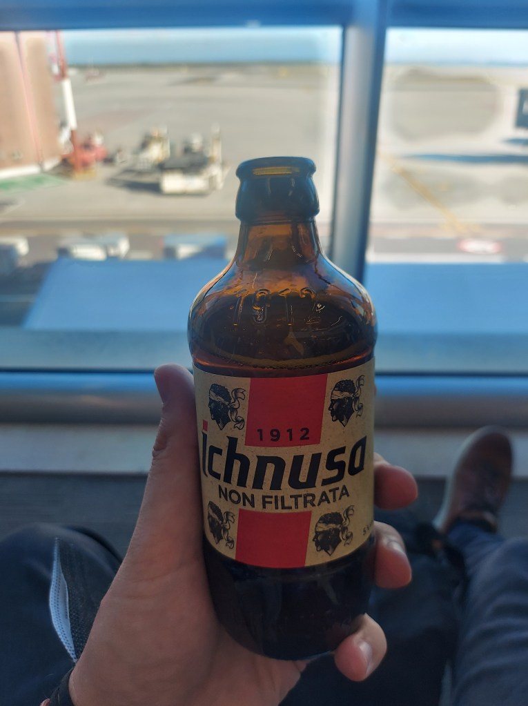 A person holding a bottle of Ichnusa beer, labeled 'NON FILTRATA', with an airport view in the background.