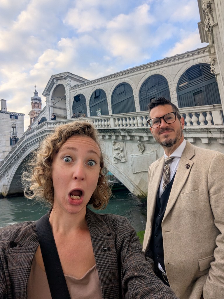 A surprised woman making a funny face in the foreground with a man in a suit smiling beside her, set against the backdrop of a historic bridge and a cloudy sky.