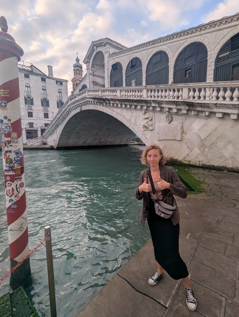 A person giving a thumbs-up gesture while standing by a canal in Venice, with the famous Rialto Bridge visible in the background.