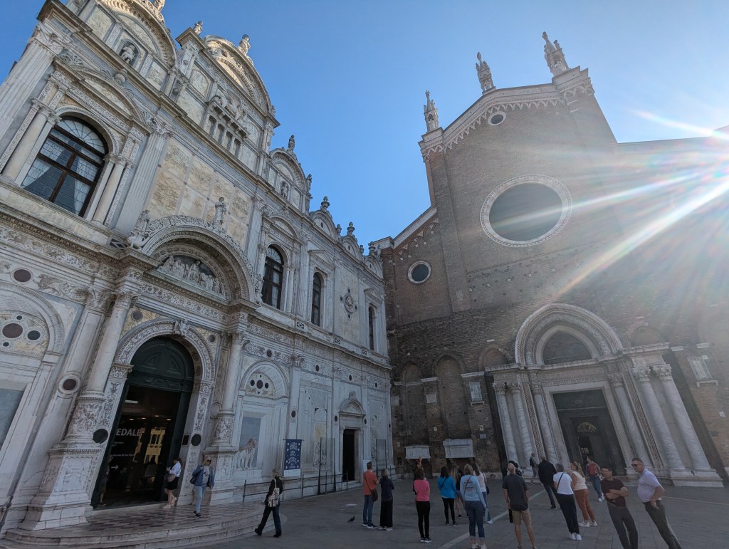 A view of two historic buildings with intricate architectural details, one featuring a stone facade and arches, and the other with a decorative entrance and artwork, set against a bright blue sky. Several people are gathered in front of the buildings.