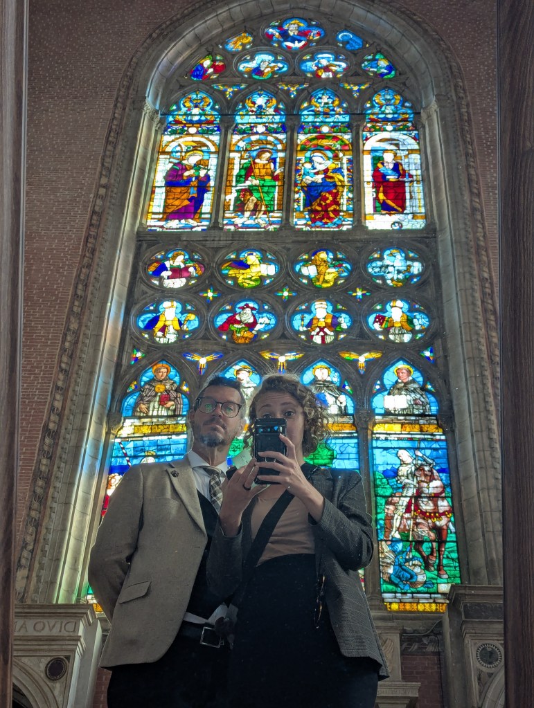 A couple posing in front of a large stained glass window inside a building, reflecting their image in a mirror.