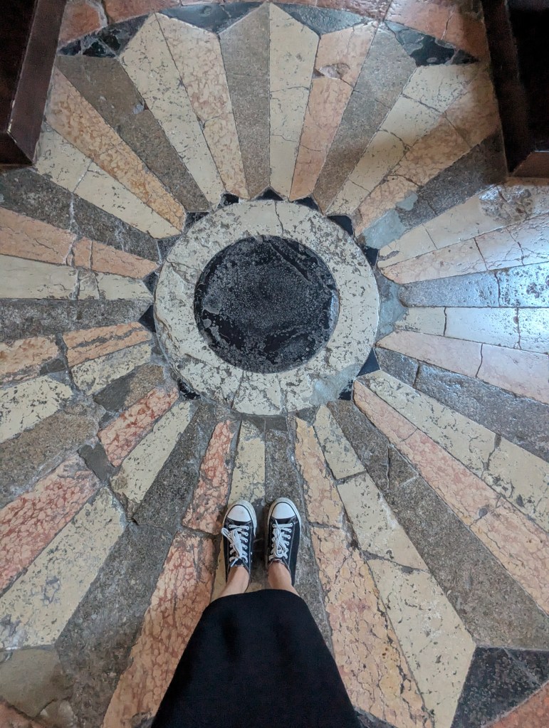 View of a decorative floor featuring a radial pattern with a central dark circle, with feet in black shoes positioned on the design.