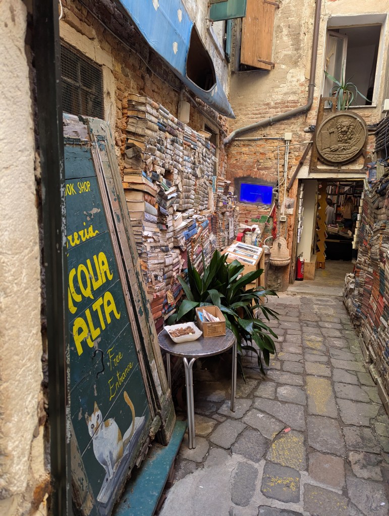 Narrow alleyway with a stacked collection of books along the wall, featuring a sign for a bookshop called 'Acqua Alta' with a painted cat. A small table displays items for sale, and there's greenery in the scene.