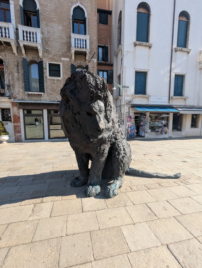 A large bronze sculpture of a sitting lion in a plaza, surrounded by historic buildings.
