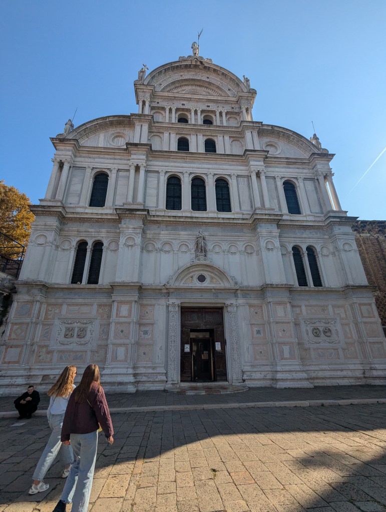 View of a historic building with ornate architecture, featuring a prominent entrance and decorative details, set against a clear blue sky. Two people are walking towards the entrance on a stone pathway.