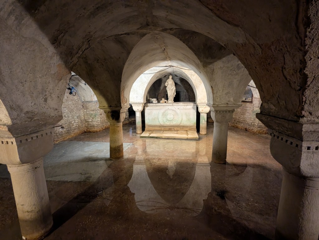 Underground chamber with arched ceilings, featuring a central stone altar and a statue, surrounded by water reflecting the architecture.