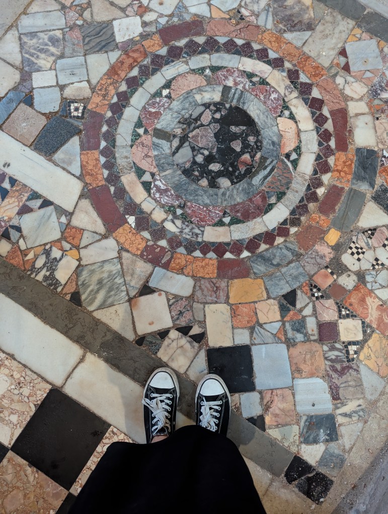 Close-up view of a colorful patterned mosaic floor featuring circular designs, with a pair of black sneakers visible in the foreground.