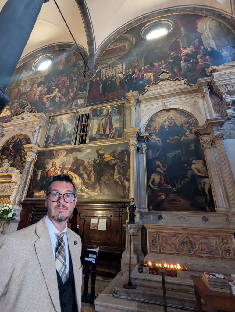 A man in a beige suit stands in a historical church with elaborate murals and paintings on the walls, including religious scenes and figures. The interior features wooden benches, candles, and a statue.