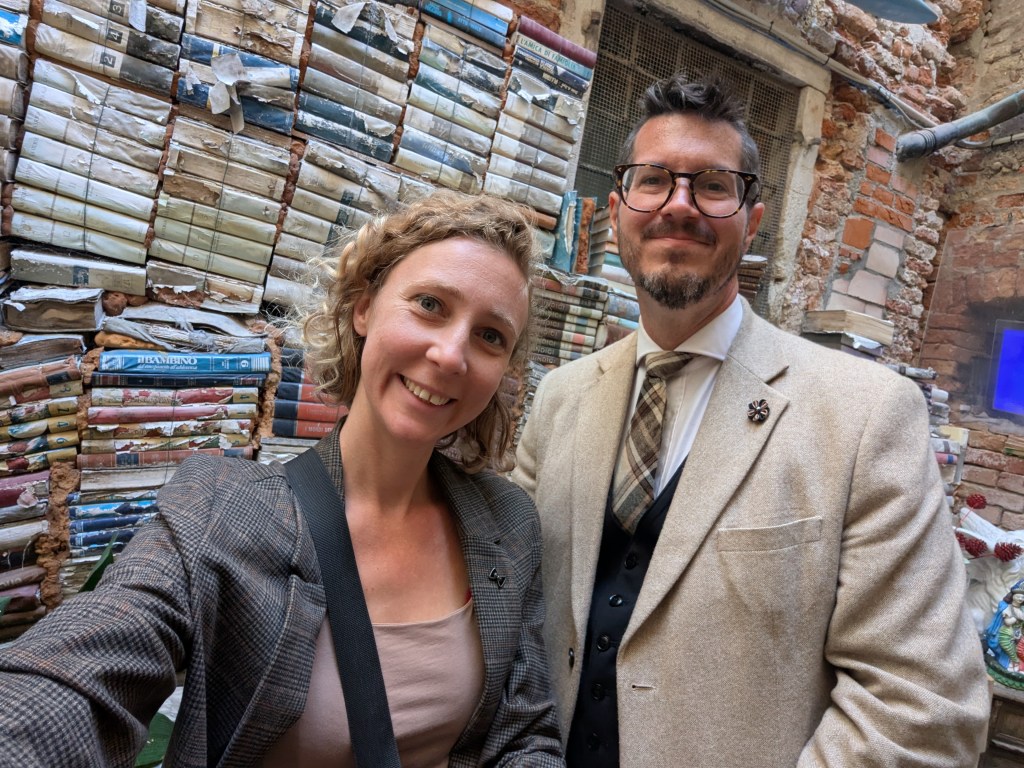A smiling woman with curly hair wearing a blazer poses for a selfie with a man in a light-colored suit and glasses, standing in front of a wall covered with stacked books.
