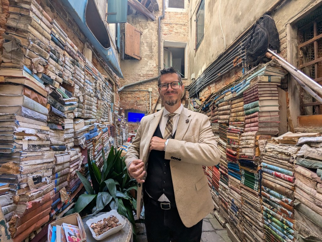 A smiling man in a beige blazer stands in a narrow alley filled with stacked books, with a green plant and some coins on a table nearby.