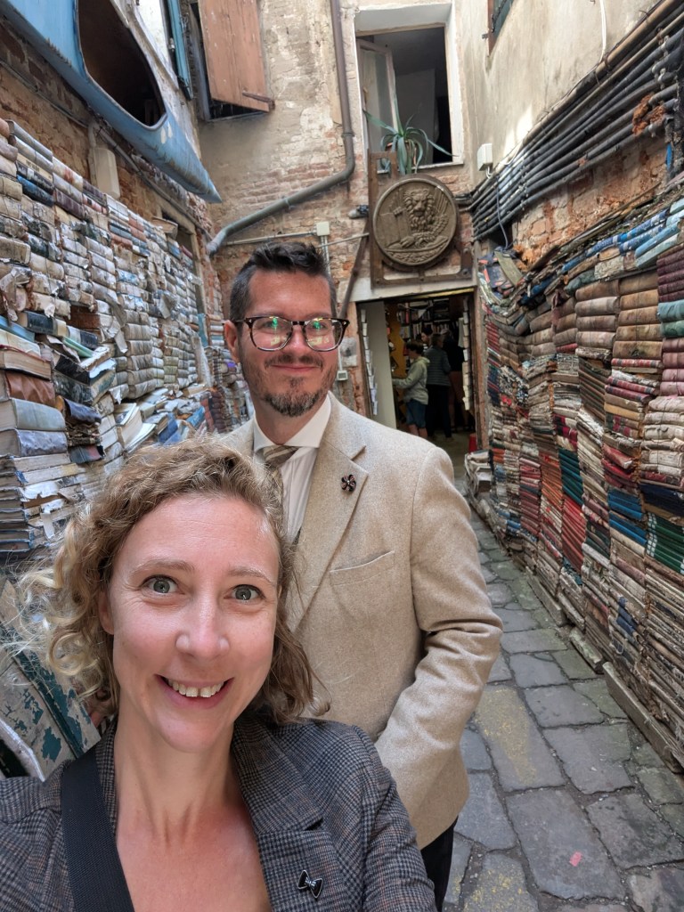 A man and a woman pose for a selfie in a narrow alley filled with stacked books. The alley features rustic brick walls and a decorative emblem in the background.