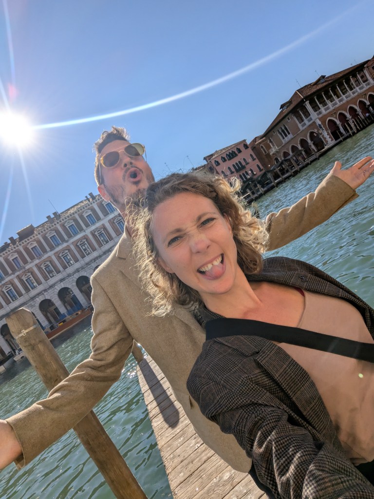 A joyful couple poses in front of a sunny waterfront in Venice, with one person playfully sticking out their tongue while the other makes an expressive pose.