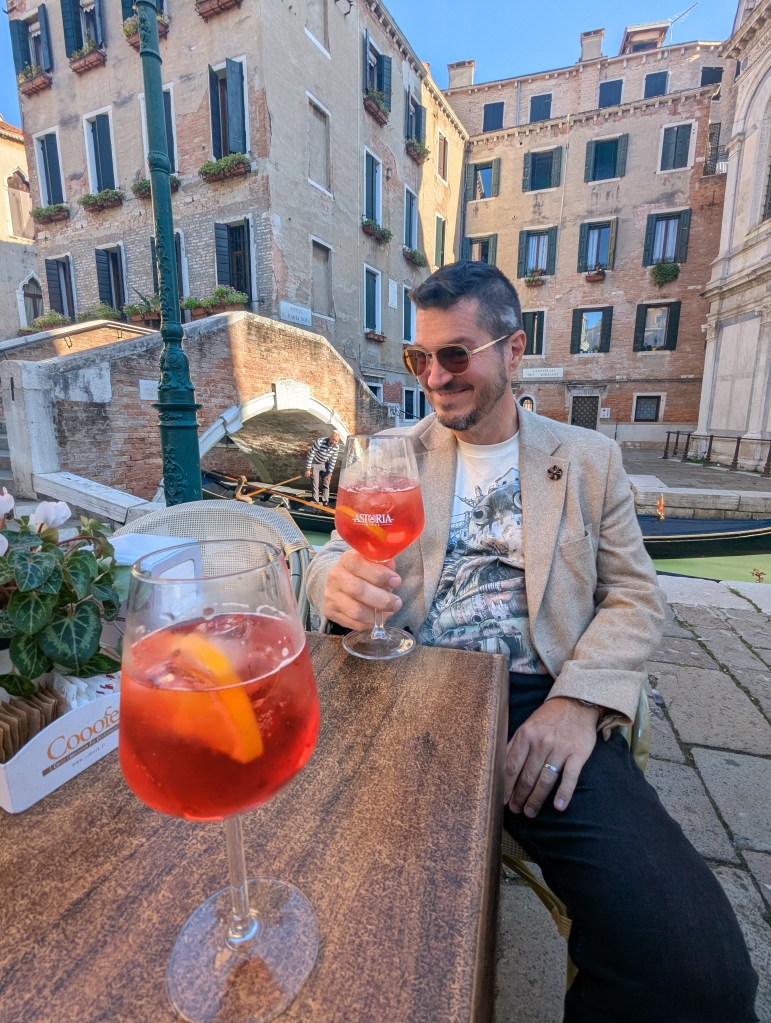A man enjoying a spritz drink at a table by a canal in Venice, with historic buildings in the background.