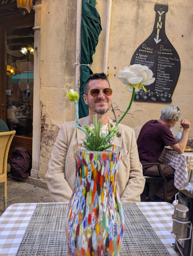 A man sitting at an outdoor café table, smiling, with a colorful vase containing a flower in the foreground. A menu board is visible in the background.