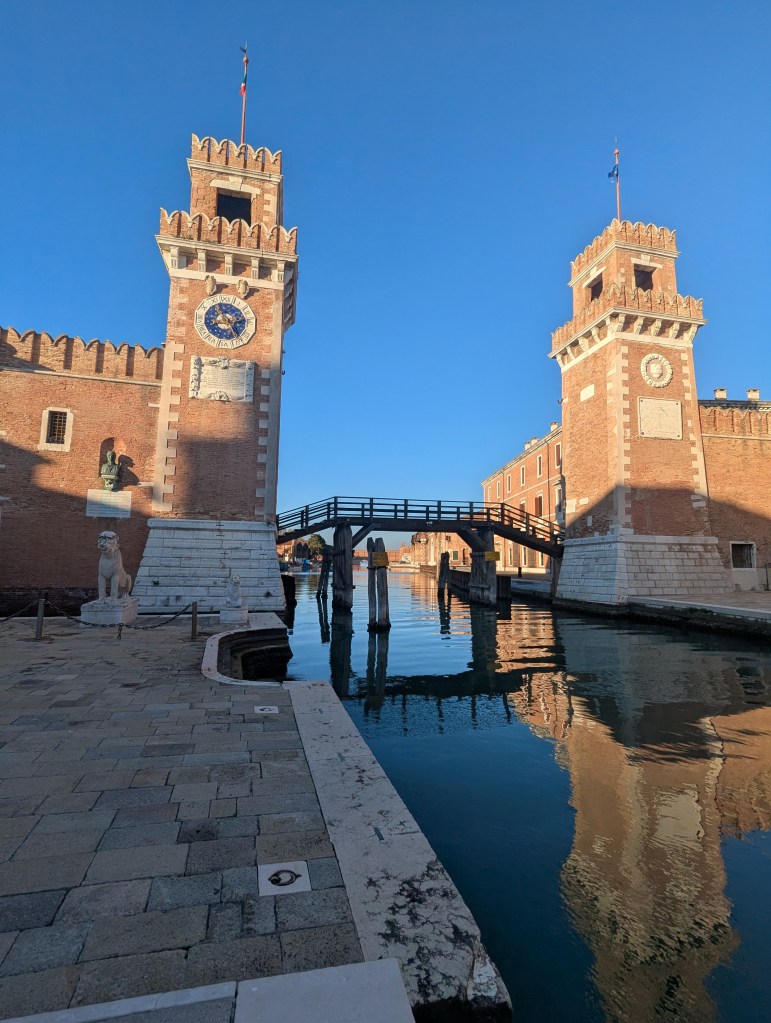 A picturesque view of two historic towers flanking a calm canal, featuring a stone bridge connecting the two sides, with clear blue skies and reflections in the water.