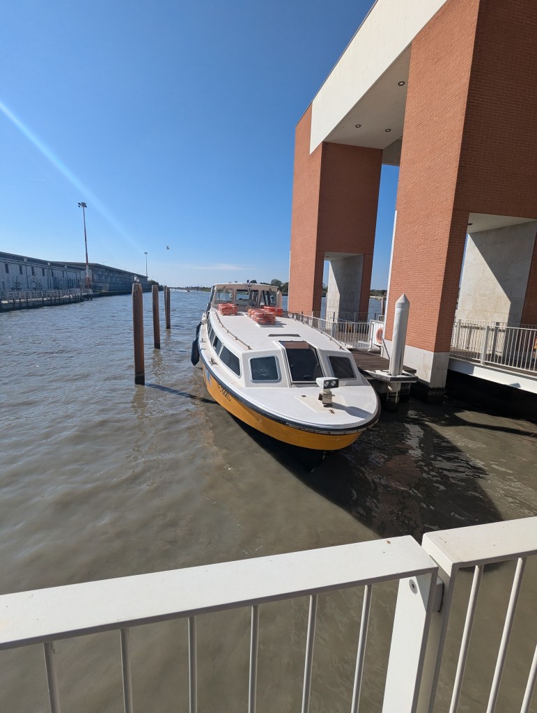 A small boat docked near a modern building, with a clear blue sky overhead and calm water surrounding it.