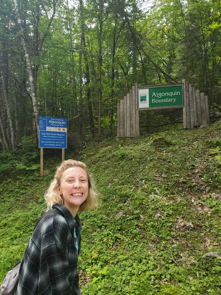 A smiling woman stands near the Algonquin Boundary sign in a wooded area, with a park information board visible in the background.