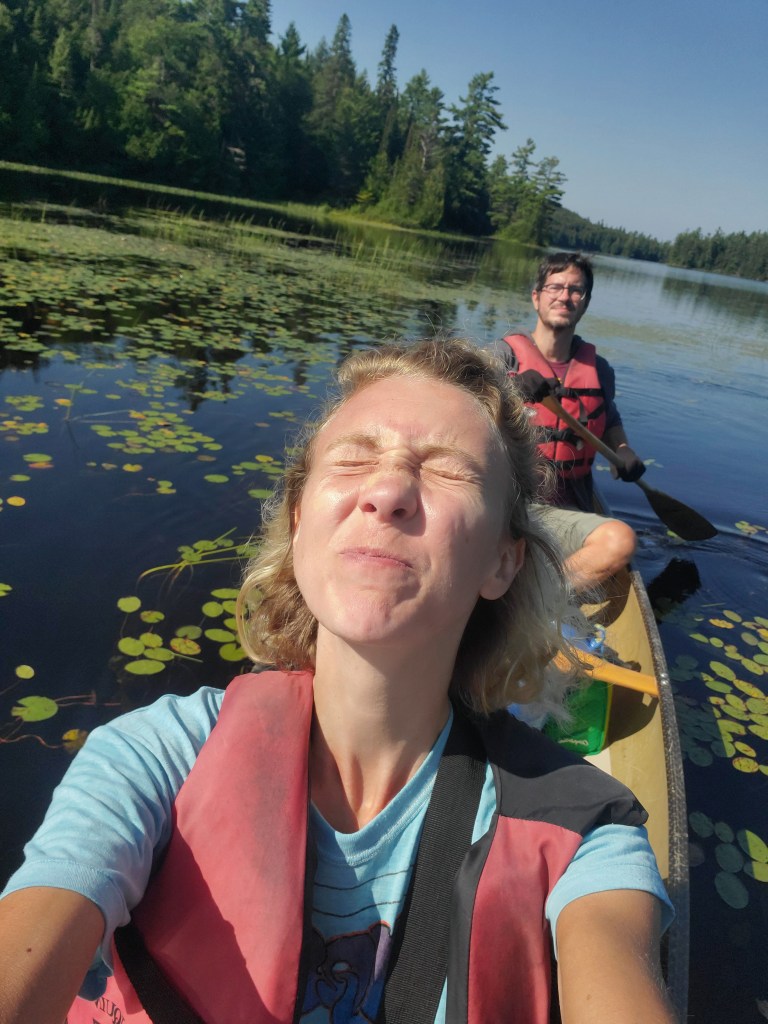 A woman enjoying the sunlight and smiling while kayaking on a calm lake, with a man paddling behind her among lily pads and lush greenery.