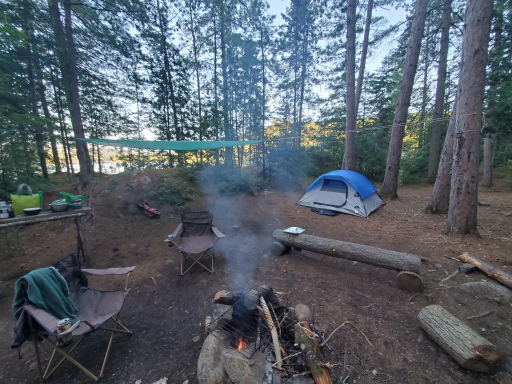 A serene camping scene featuring a blue tent, a campfire with smoke, two folding chairs, and a shaded cooking area surrounded by tall trees.