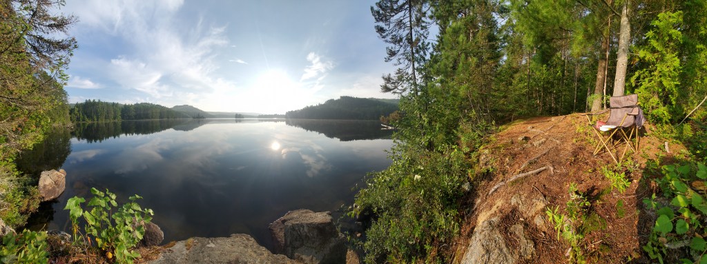 A panoramic view of a tranquil lake surrounded by lush greenery and trees, with a portable chair positioned on the rocky shore, reflecting the calm water and bright sunlight.