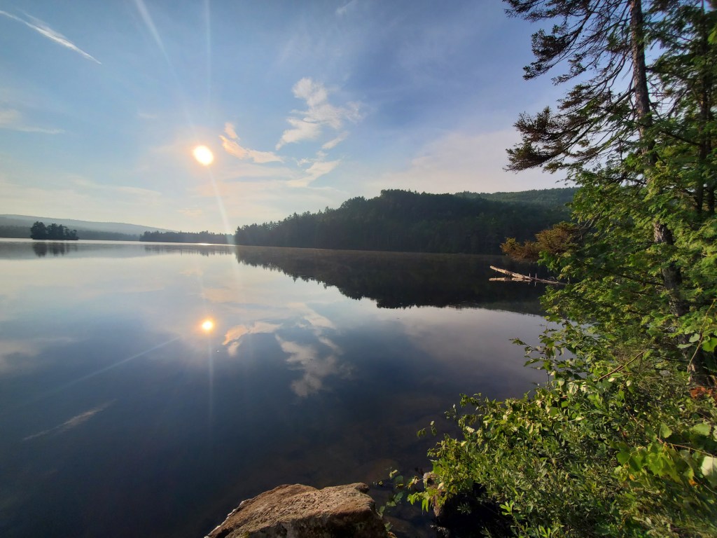 Scenic lake view at sunrise with reflections in the calm water, surrounded by trees and hills.