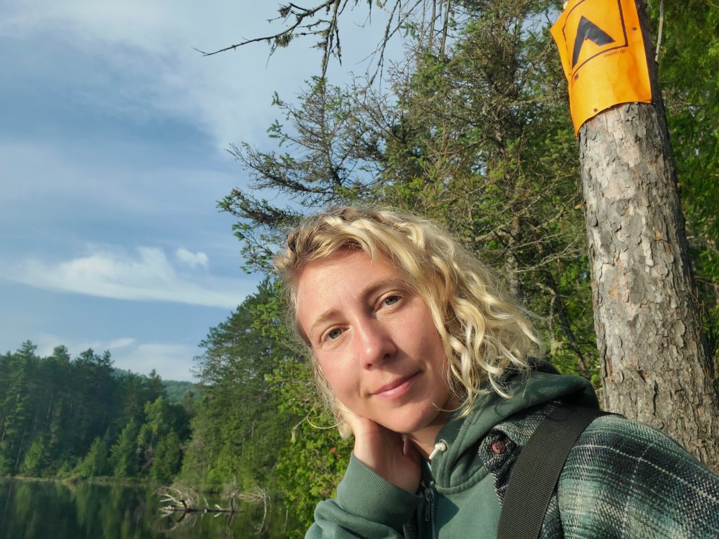 A person with curly hair standing near a lake surrounded by trees, with a yellow marker on a tree in the background.