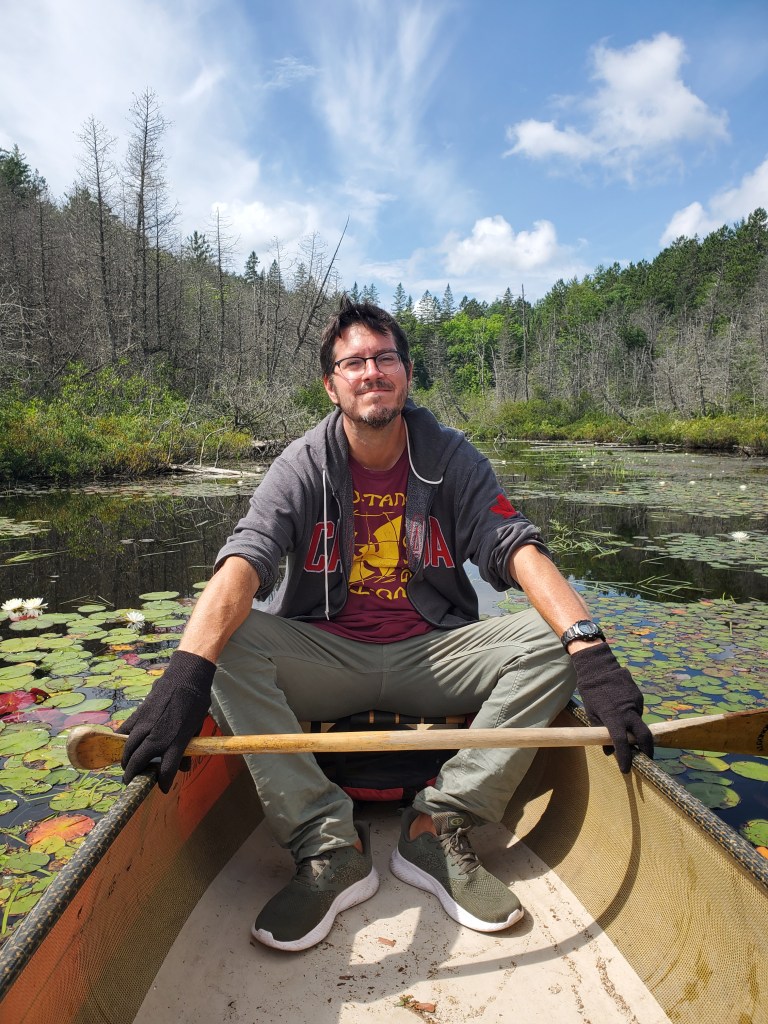 A person sitting in a canoe on a serene body of water, surrounded by lily pads and greenery, smiling and holding a paddle, wearing casual clothing and gloves, with a clear blue sky overhead.