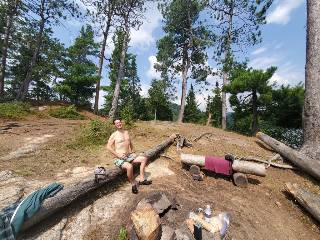 A person sitting on a log at a campsite surrounded by trees and a lake in the background. The individual is shirtless, wearing colorful swim trunks, and appears to be enjoying the outdoors.