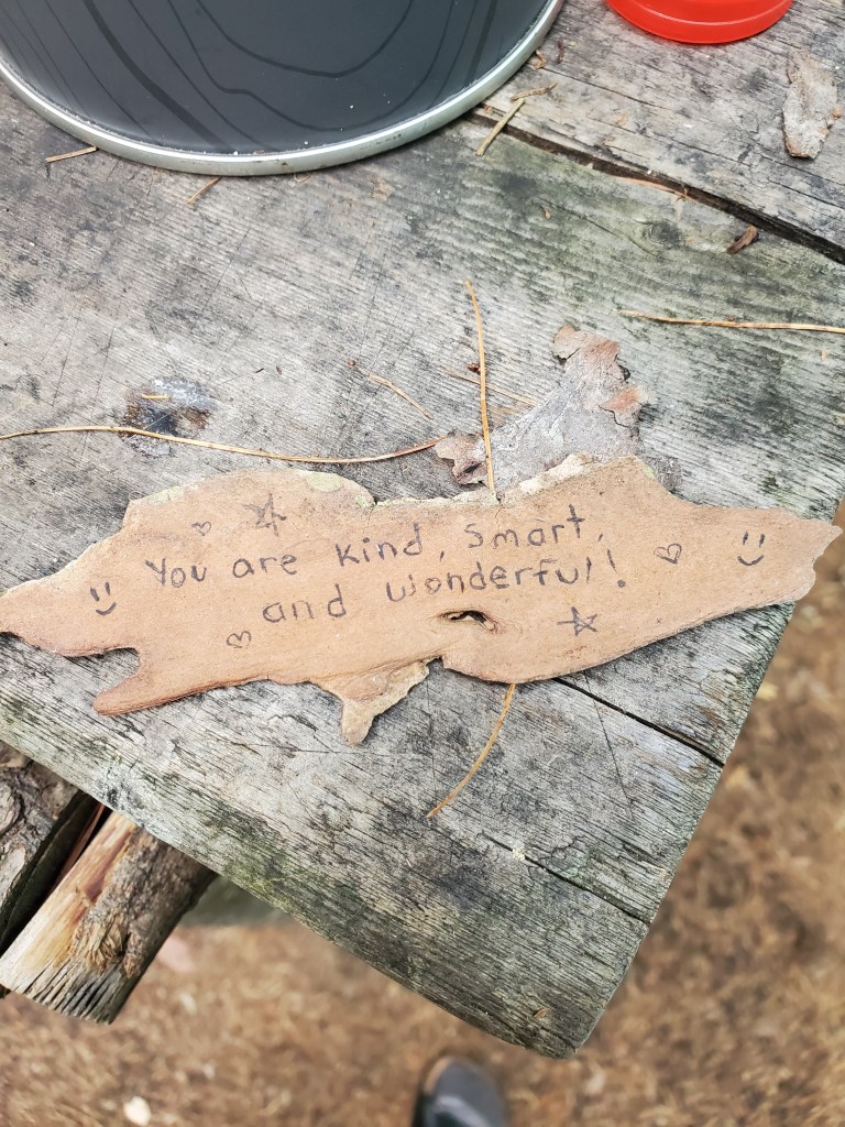 A piece of bark with a handwritten message that reads 'You are kind, smart, and wonderful!' surrounded by small drawings of hearts and smiley faces, placed on a wooden surface.