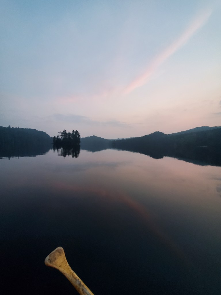 A calm lake at dusk, reflecting the surrounding mountains and a soft pink sky, with a canoe paddle partially visible in the foreground.