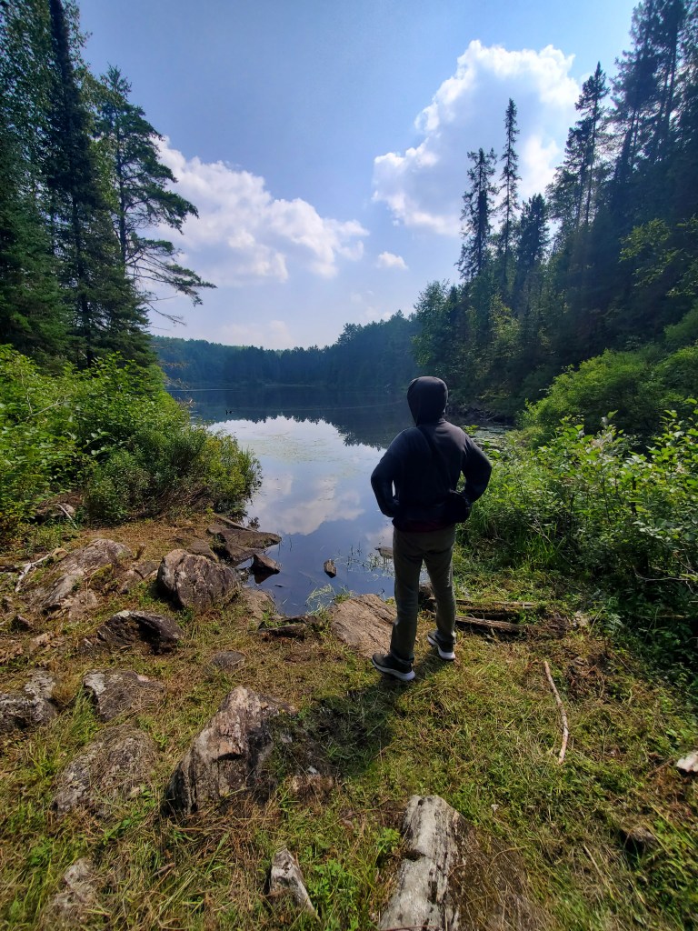 A person standing on the rocky shore of a lake, surrounded by lush greenery and trees, with clouds in a blue sky above.