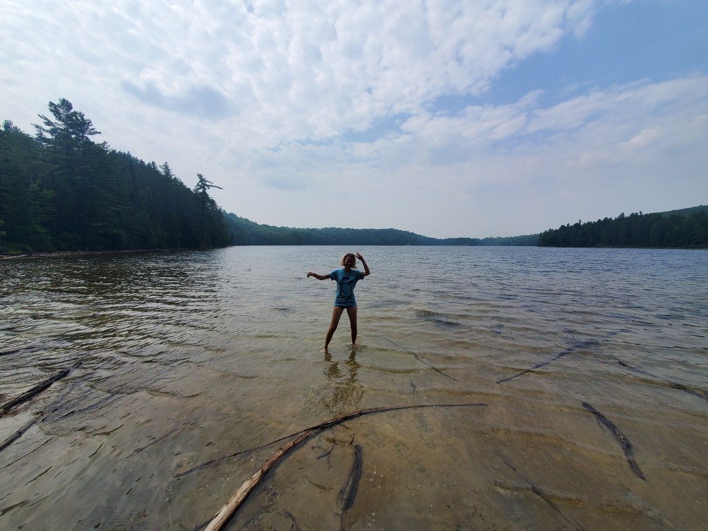 A person standing in shallow water at a serene lake, surrounded by trees and a partly cloudy sky.