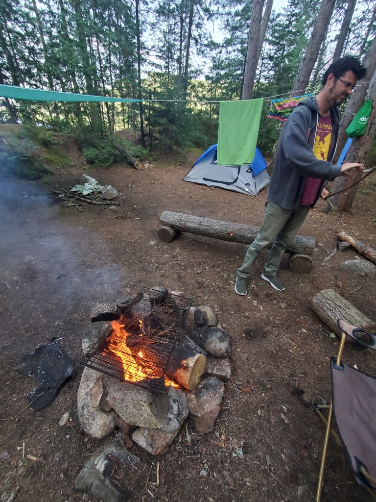 A campsite scene featuring a campfire with flames and smoke, surrounded by a stone circle. In the background, a blue tent is visible, along with clothes hanging on a line between trees. A person stands nearby, manipulating the fire.