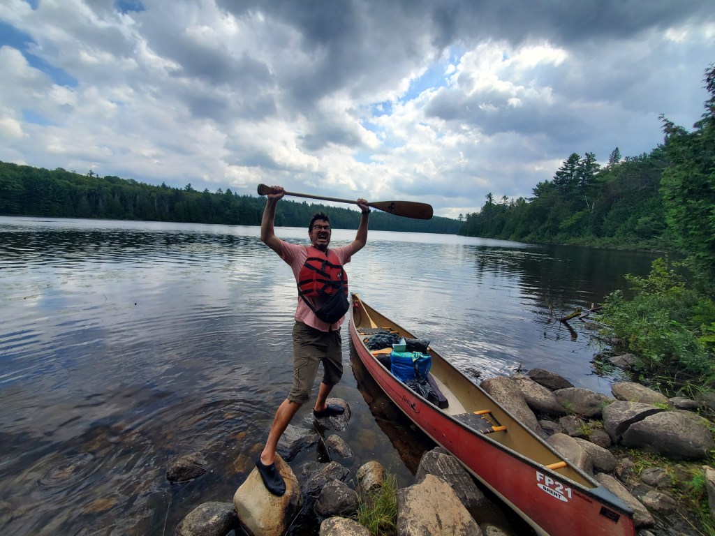 Person standing on rocks by a lake, playfully raising a canoe paddle with a canoe partially visible in the water.