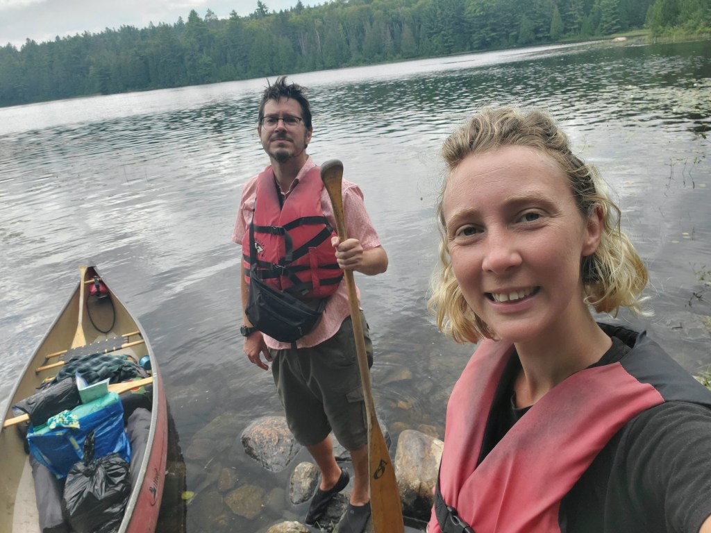 A man and a woman standing on a rocky shore beside a canoe at a calm lake. They are wearing life jackets and smiling for the camera, with lush trees in the background.