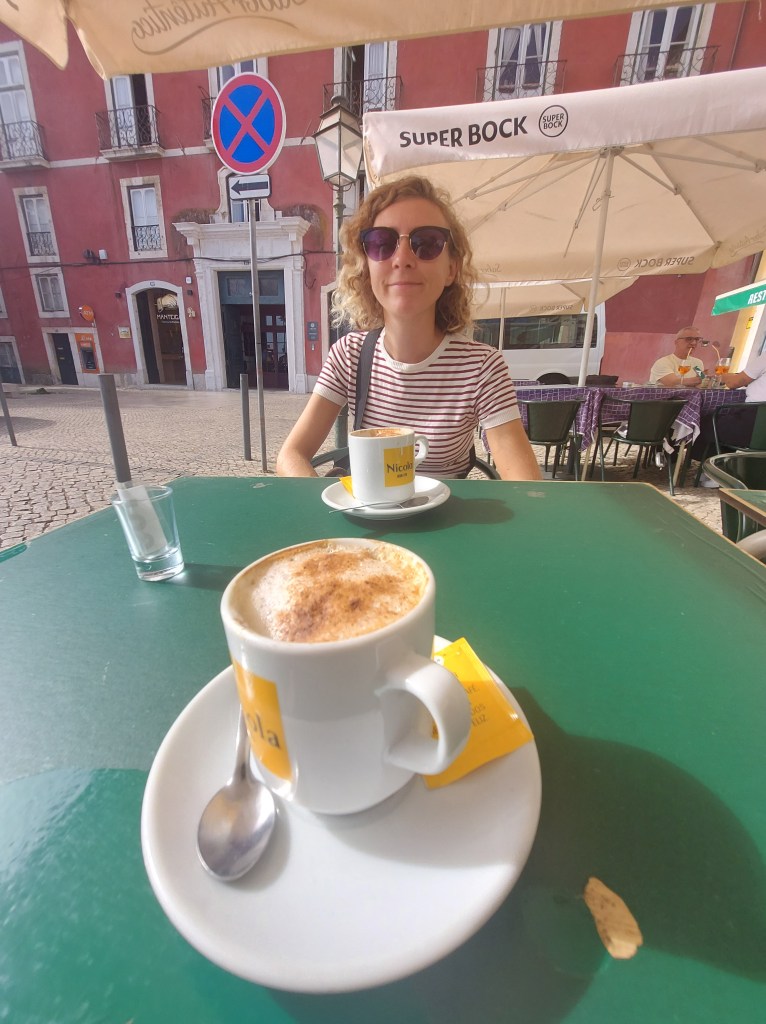 A woman sitting at an outdoor cafe table with a cup of coffee in front of her. The scene includes a green table, a street view with a no-parking sign, and umbrellas, indicating a sunny day.