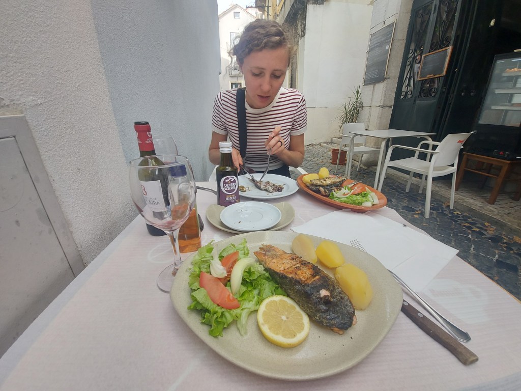A woman sitting at an outdoor table enjoying a meal of grilled fish with potatoes and salad on a sunny day, with a bottle of wine and a glass nearby.