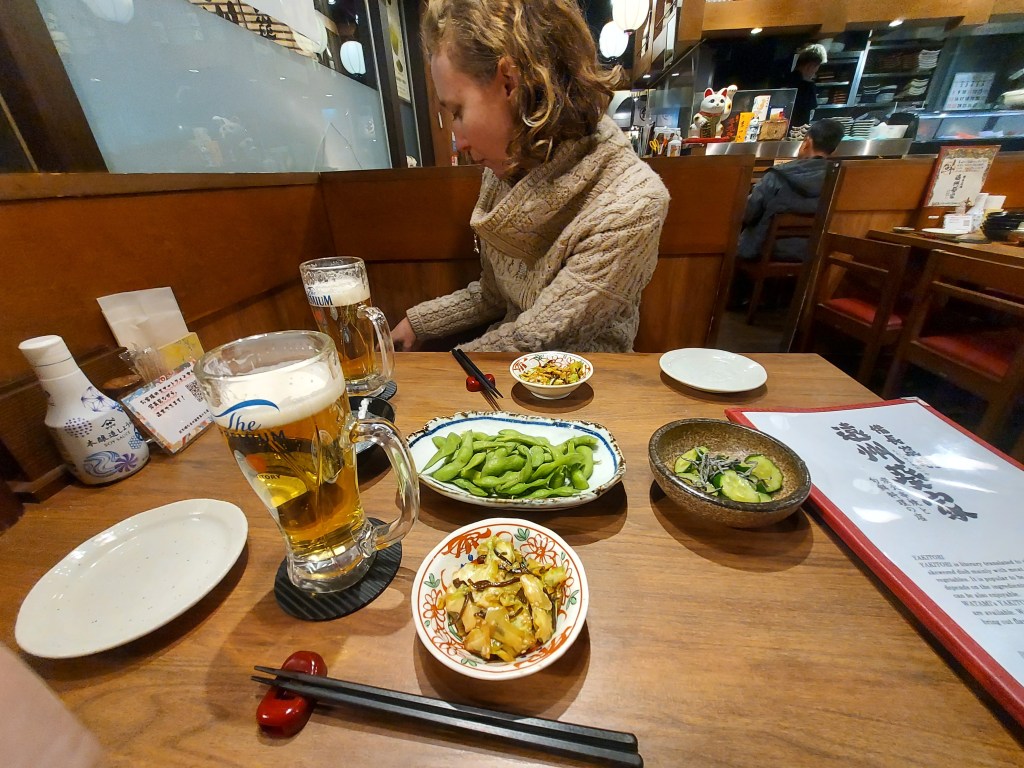 A cozy dining table at a Japanese restaurant featuring two mugs of beer, a selection of side dishes including edamame and assorted vegetables, chopsticks, and a menu on the table. A person sits at the table, partially visible.