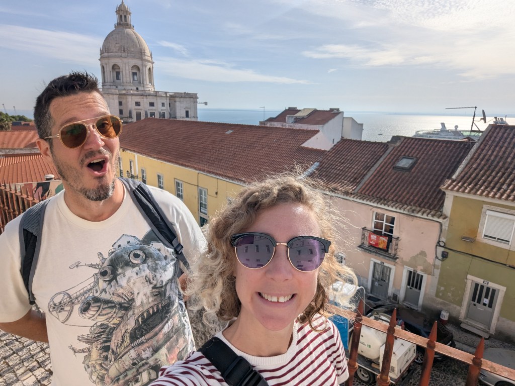 A man and a woman taking a selfie with a scenic view of rooftops and the ocean in the background. The man has sunglasses and a surprised expression, while the woman smiles and wears sunglasses. A dome building is visible in the background.
