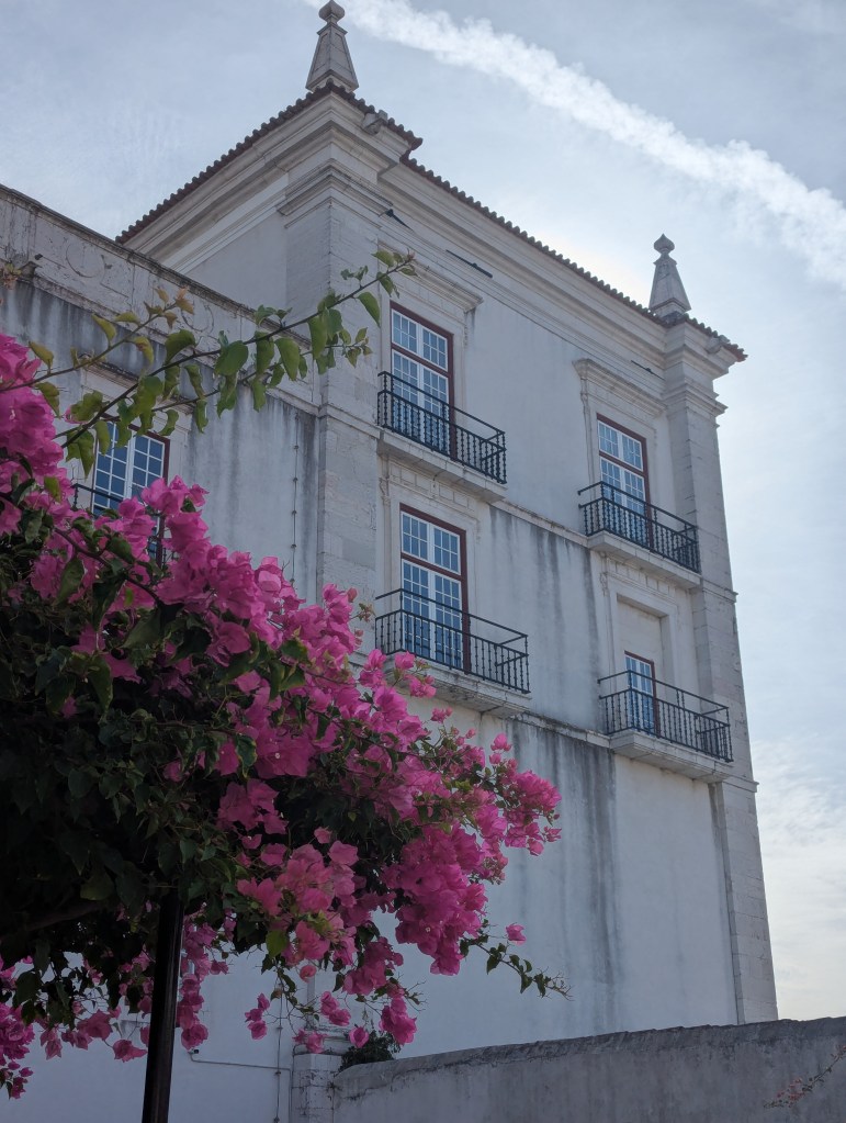 A historical building with white walls and decorative features, featuring balconies and a pointed roof, framed by vibrant pink bougainvillea flowers in the foreground.