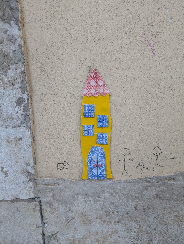 A playful street artwork featuring a colorful paper house with a red roof and blue windows attached to a textured wall, accompanied by simple stick figure drawings nearby.
