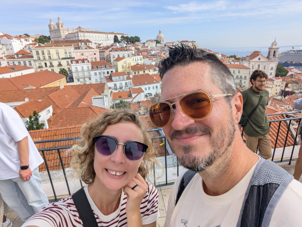 A couple takes a selfie with a scenic view of a city featuring red rooftops, churches, and the ocean in the background.