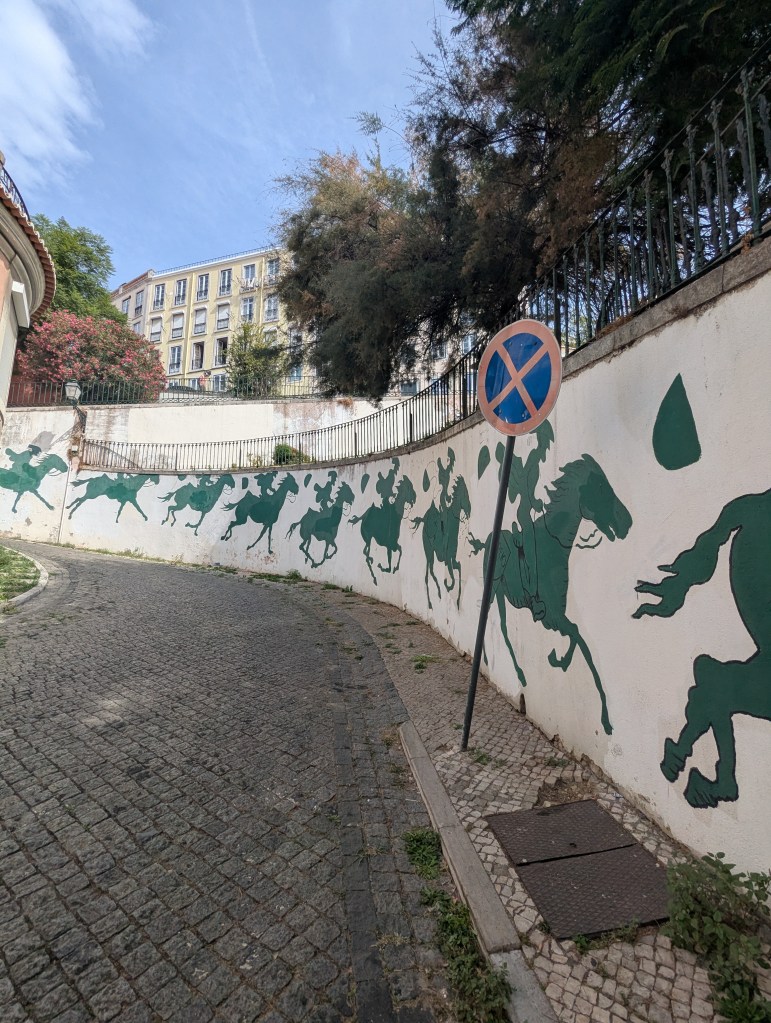 A curved street with a mural depicting green horse riders on a white wall, alongside a no parking sign.
