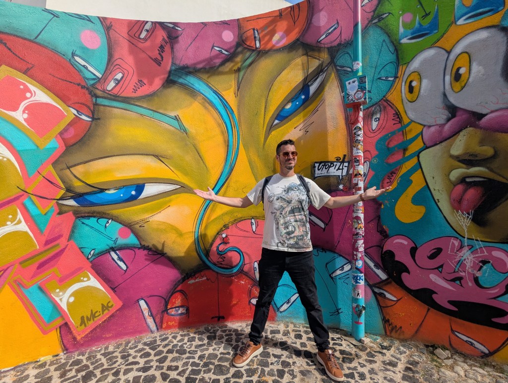 A person stands with arms outstretched in front of a vibrant urban mural featuring colorful abstract faces and eyes.