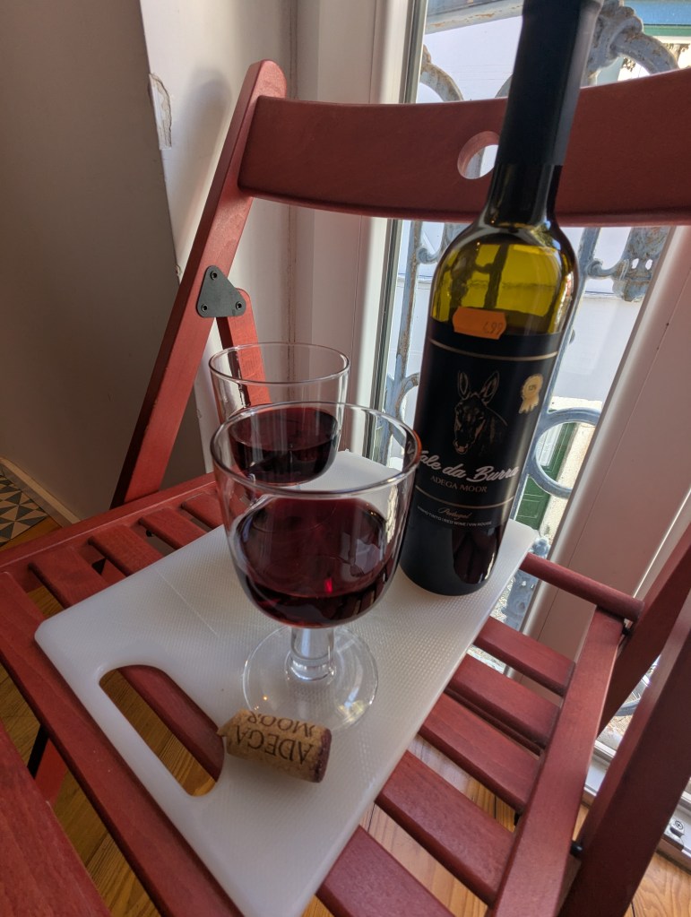 A bottle of red wine labeled 'Adega Mook' next to two glasses of wine on a white cutting board, placed on a red wooden chair near a window.