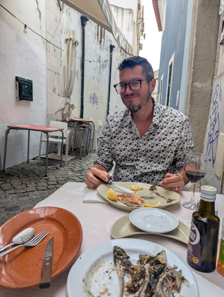 A man sitting at a table in an outdoor alleyway, enjoying a meal with plates showing remnants of fish. He is smiling and holding a fork and knife, with a glass of red wine and olive oil on the table.